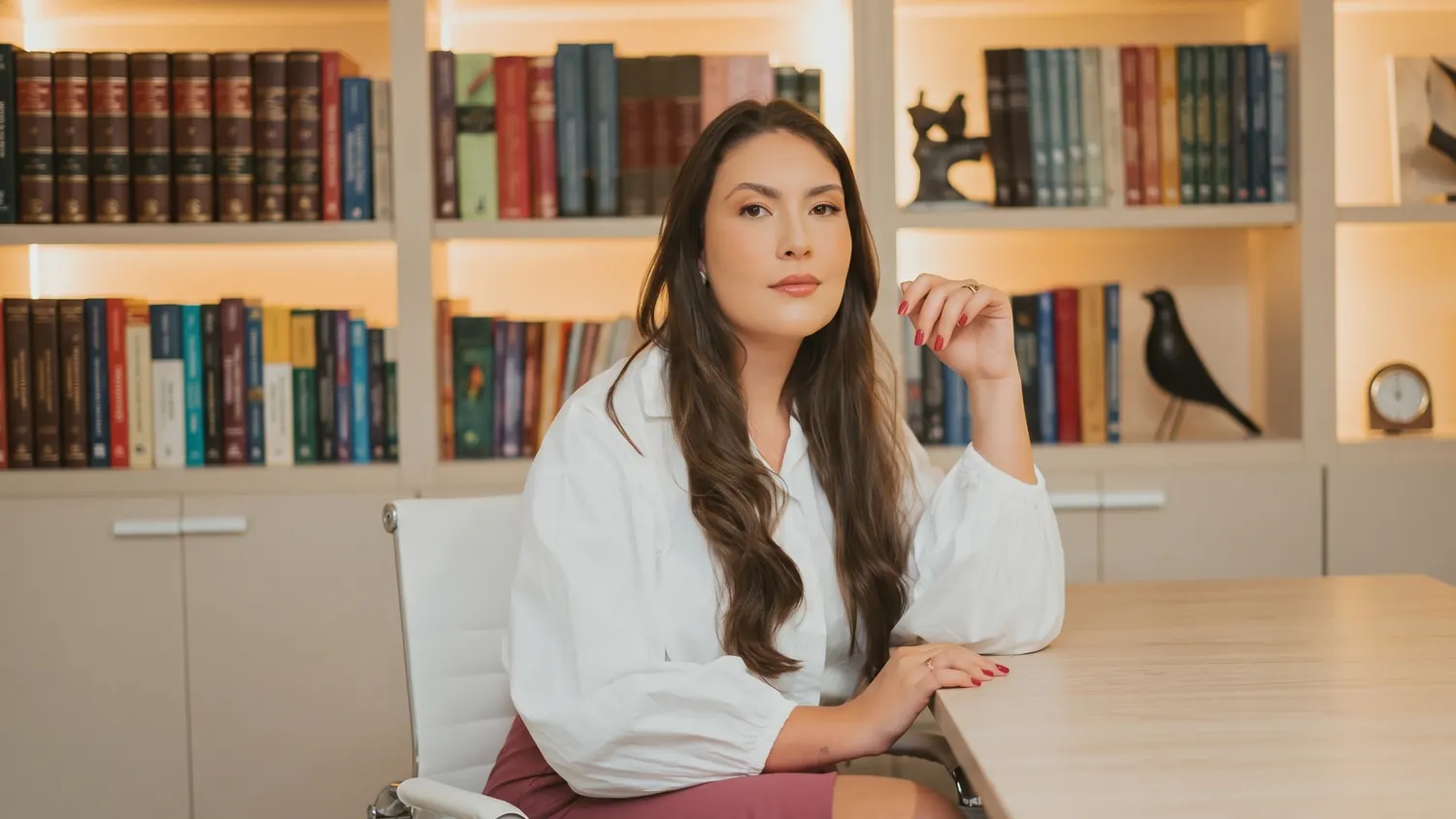 Lorena Galli sentada à mesa do escritório, camisa branca, estante de livros jurídicos ao fundo